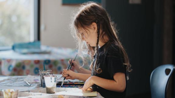 A young girl happily creating art at a table, immersed in her painting with a variety of colors and tools nearby.