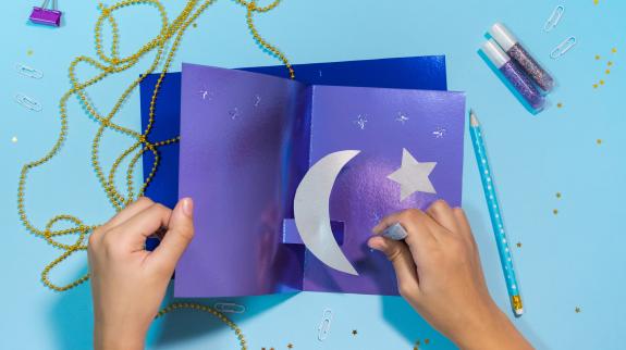 An overhead view of a pair of hands working on a purple and silver Ramadan craft. String, clips, and glitter glue sit on the table around the craft.