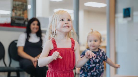 Two toddler girls try to catch bubbles at Toddler Time while their parent watches in the background.