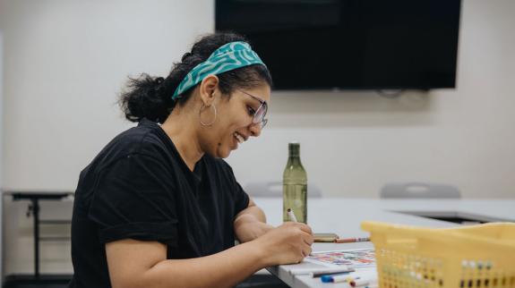 South Asian woman (30s) with teal headband sits at table surrounded by art supplies while she colours a sheet.