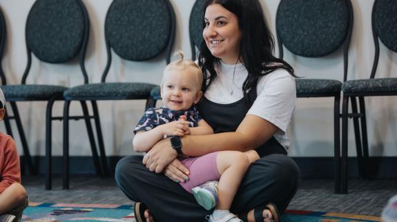 A Caucasian mom (30s) sits on floor with toddler girl in her lap at interactive library program.