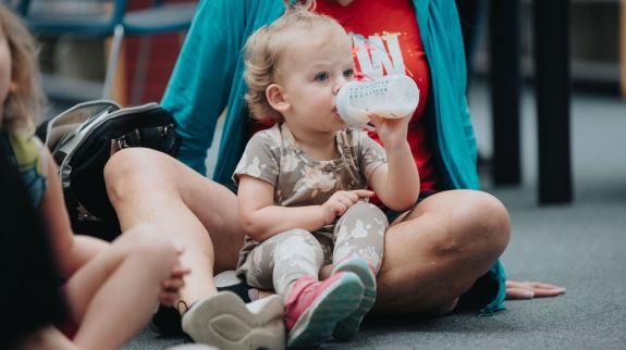 A mom and baby girl sit on the library floor together listening to stories while the baby drinks from a bottle.