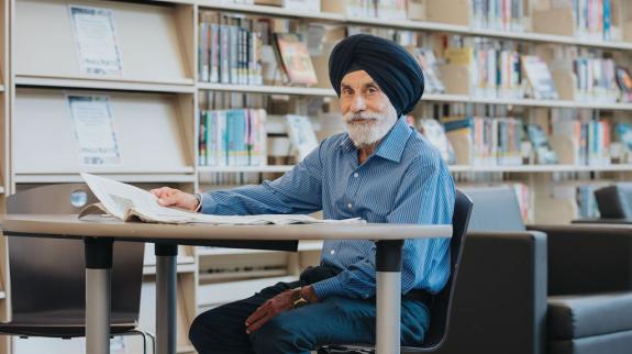 A senior South Asian man (70s) looking up sits at table with a book at the library.