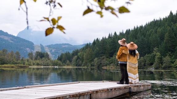 Two Indigenous people stand at the end of a dock looking out at the lake.