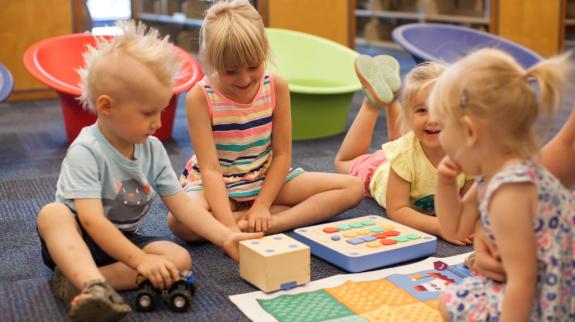 A group of children engaged in play, learning to code with Cubetto through stories and hands-on play, in a bright and inviting library space.