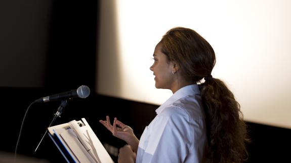 A female (20s) stands in front of a microphone, reading a poem to the unseen audience in front of her.