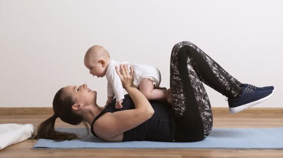 A mom, working out laying on her back, holds her infant above her. She bonds with her infant while doing physical activity.