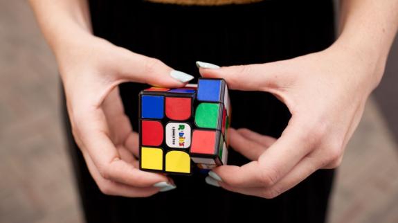 A pair of hands at waist level, with white painted nails, work to solve a Puzzle Cube.