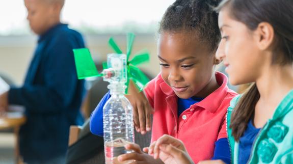 African American and Hispanic elementary age little girls are building a windmill from a water bottle during an after school science program in a public library. Students are learning science, technology, engineering, and math while working on a project together.