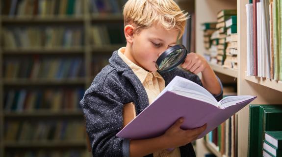 A boy uses a magnifying glass to spy things in a book from the library shelf.