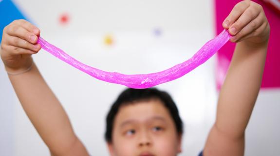 A young boy (8) holds up stretched out magenta coloured slime above his head.