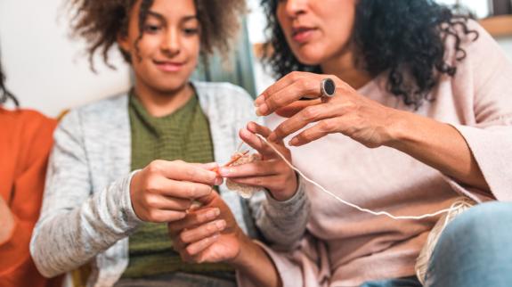 An older woman teaches a teenage girl basic knitting stitches in a relaxed, homey environment.