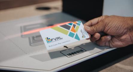 A person holds a library card near a machine, indicating they are about to use it for a payment or identification process.