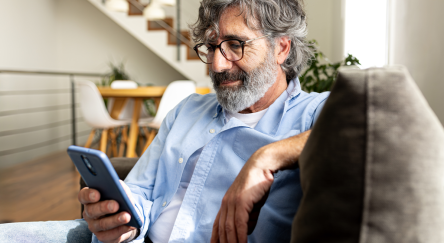 A man with glasses and a beard sits comfortably on a couch, intently looking at his phone.