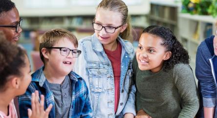 A multi-ethnic group of six elementary school students standing together in the library. They are 10 and 11 years old. The boy in the plaid shirt, wearing eyeglasses, has down syndrome.