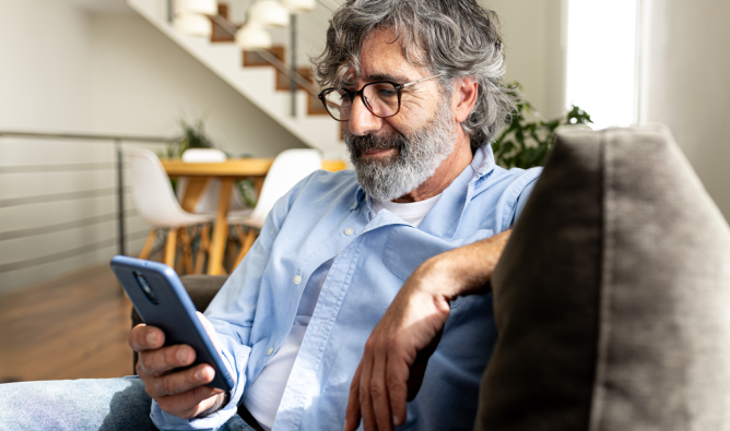 A man with glasses and a beard sits comfortably on a couch, intently looking at his phone.