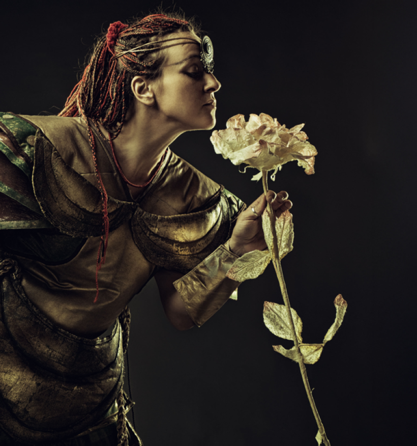 A woman clad in armor delicately holds a colorful flower, blending strength and beauty.